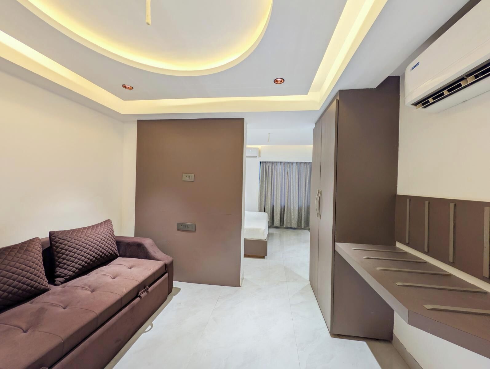 A modern hotel room with a sofa, a bed, a wardrobe, and a ceiling with recessed lighting.