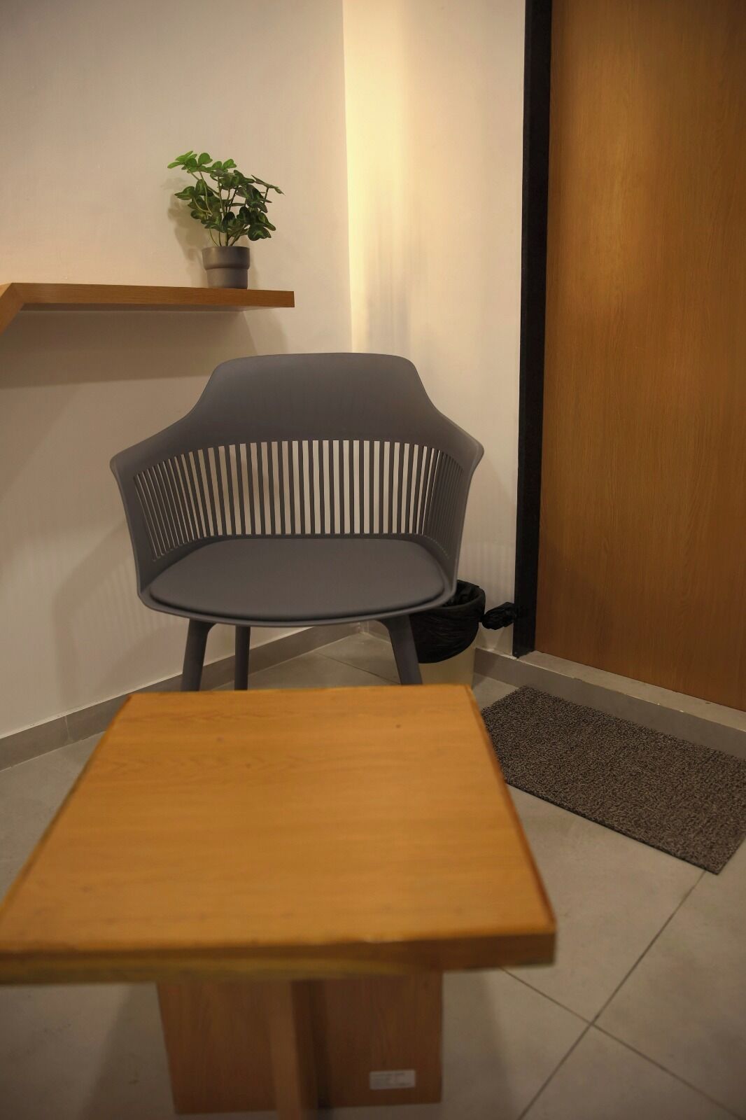 Um quarto pequeno com uma mesa de madeira, uma cadeira estofada cinza, uma planta em vaso sobre uma estante e uma porta de madeira.