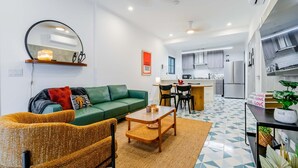 Una sala de estar moderna con un sofá verde, una mesa de centro y un comedor con sillas. Se observa una cocina al fondo.
