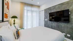 Una habitación de hotel moderna con una cama grande, un ventilador de techo, un televisor montado y una ventana con cortinas translúcidas.