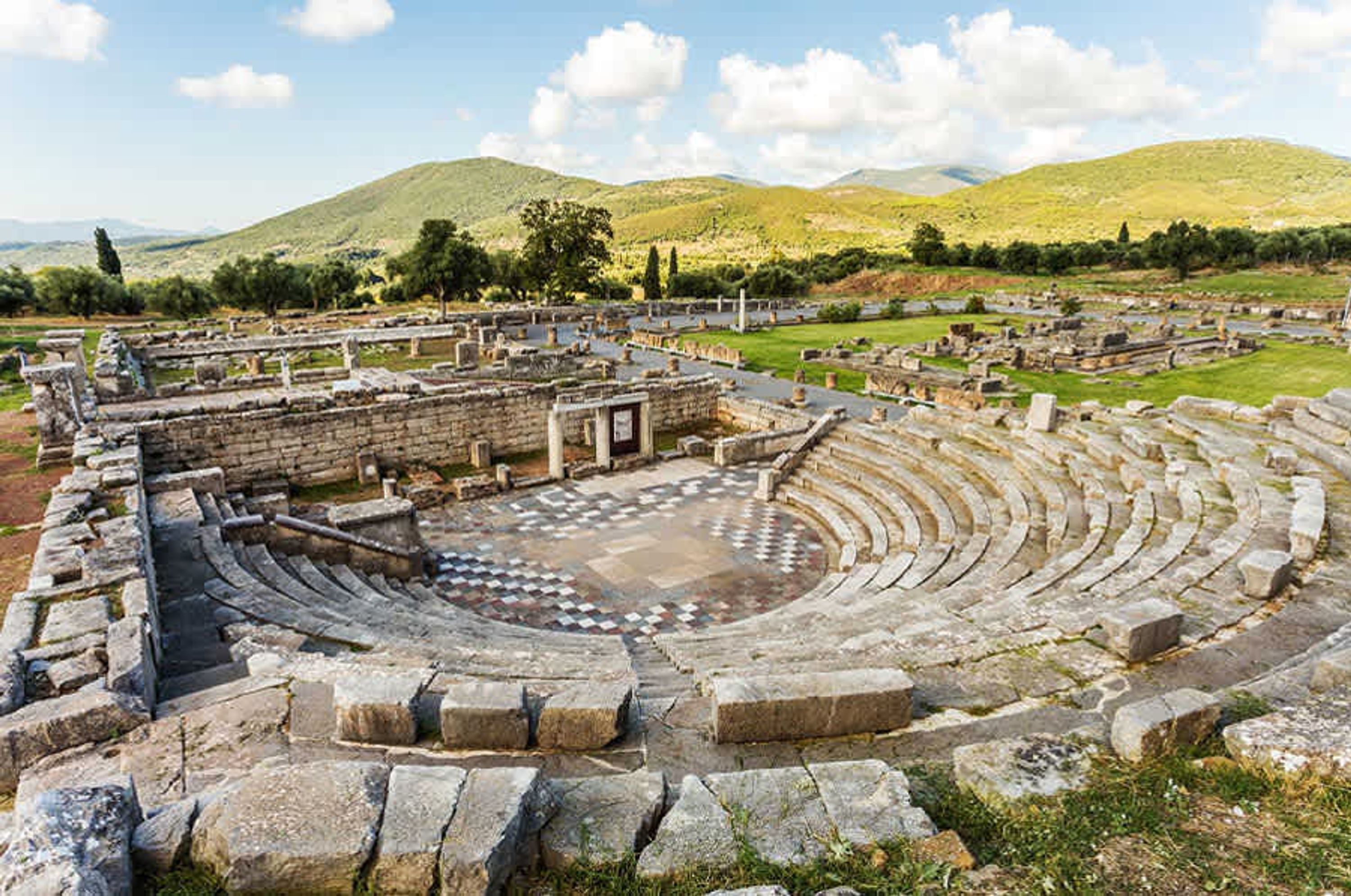 An ancient amphitheater with stone seating and a central stage, surrounded by ruins and hills.