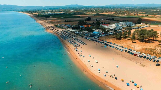 Vista aérea de una playa con hileras de sombrillas, un estacionamiento y edificios al fondo.