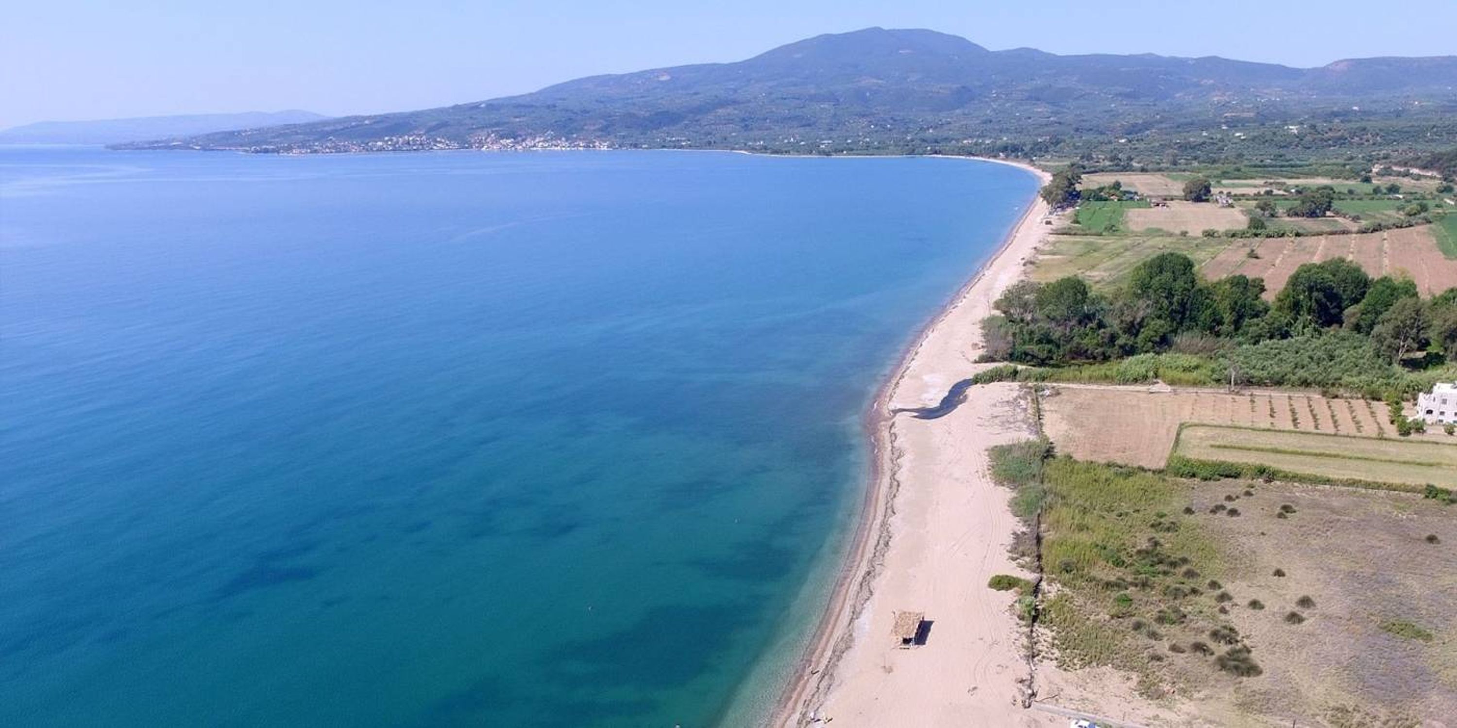 Vue aérienne d’une zone côtière avec une plage, des champs et une montagne en arrière-plan.
