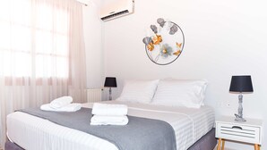 Una cama bien hecha con ropa de cama blanca y gris, un aire acondicionado de pared, una obra de arte decorativa en la pared y una mesita de noche con una lámpara.