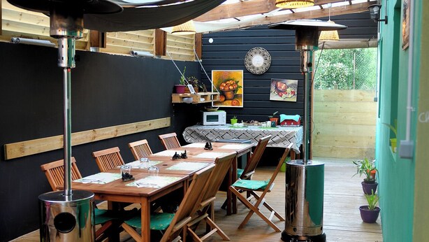 An outdoor dining area with a long wooden table, chairs, and a metal trash can.