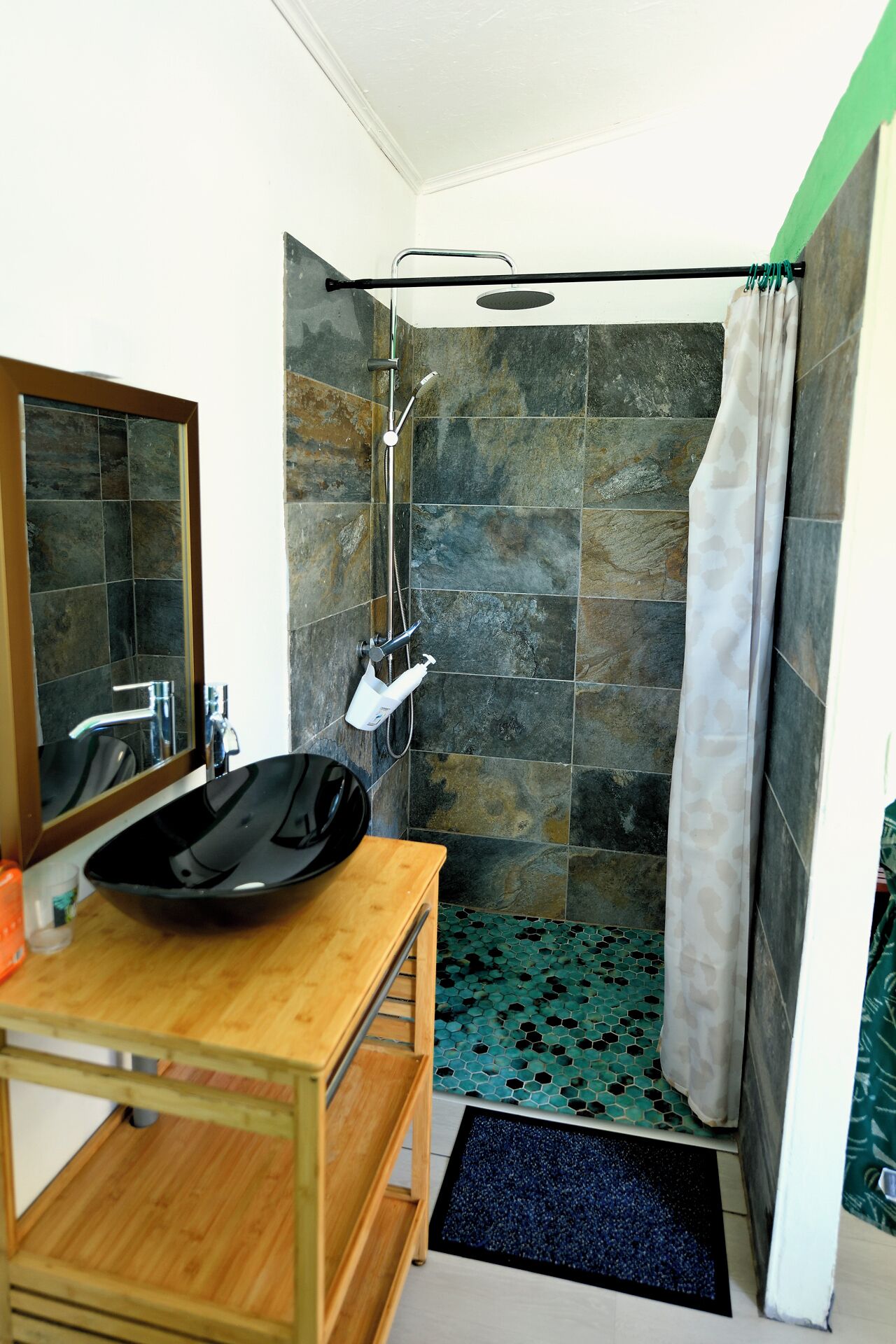 Casa de banho com lavatório preto, espelho, chuveiro com chão padronado em verde e preto, e cortina de chuveiro.
