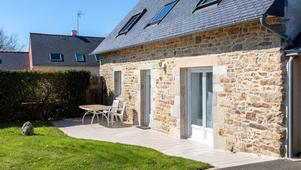 A stone building with a tiled roof, a patio with a table and chairs, and a hedge in the background.