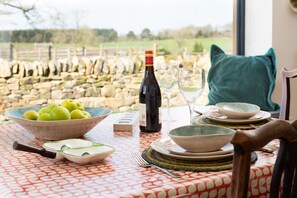 A table set with a bowl of green apples, a bottle of wine, and wine glasses, with a stone wall and sheep in the background.