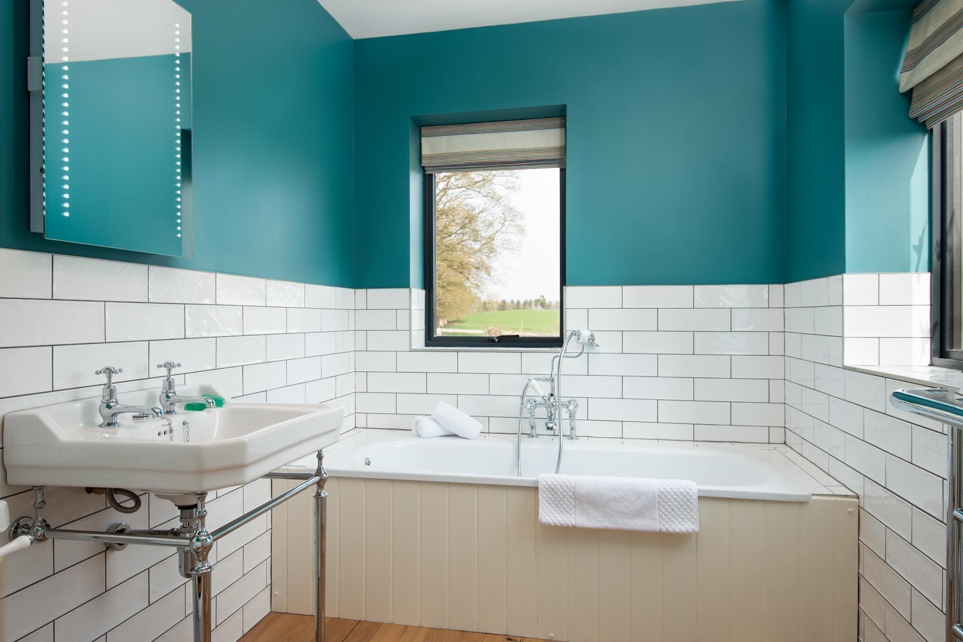 A modern bathroom with teal walls, white tiles, a bathtub, and a window with a view of greenery.
