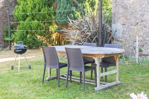 An outdoor dining area with a wooden table and wicker chairs, surrounded by greenery and a stone wall.