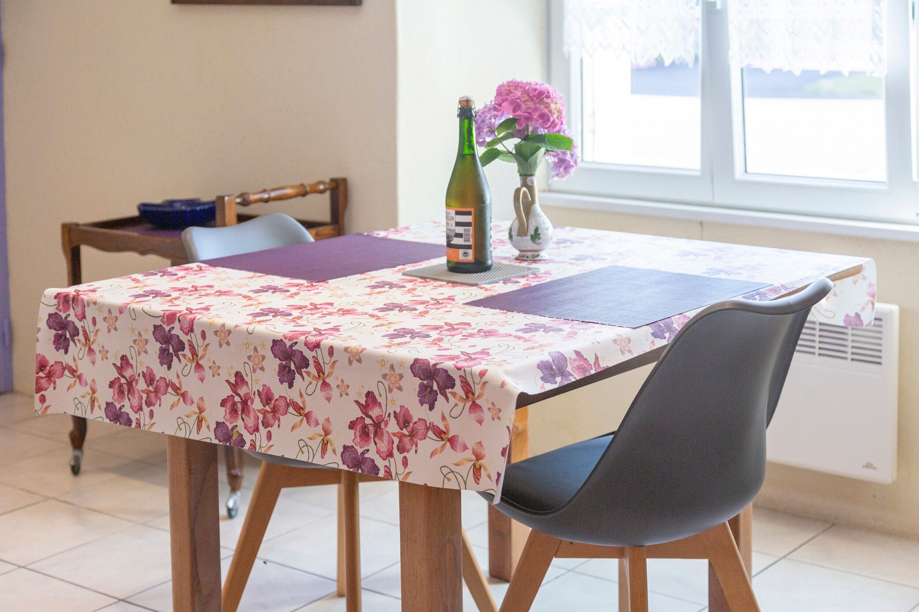 Une table à manger recouverte d’une nappe à motifs floraux, une bouteille de vin et un vase rempli de fleurs.