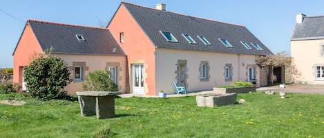 A two-story house with a sloped roof, a small garden with a stone bench, and a blue chair.
