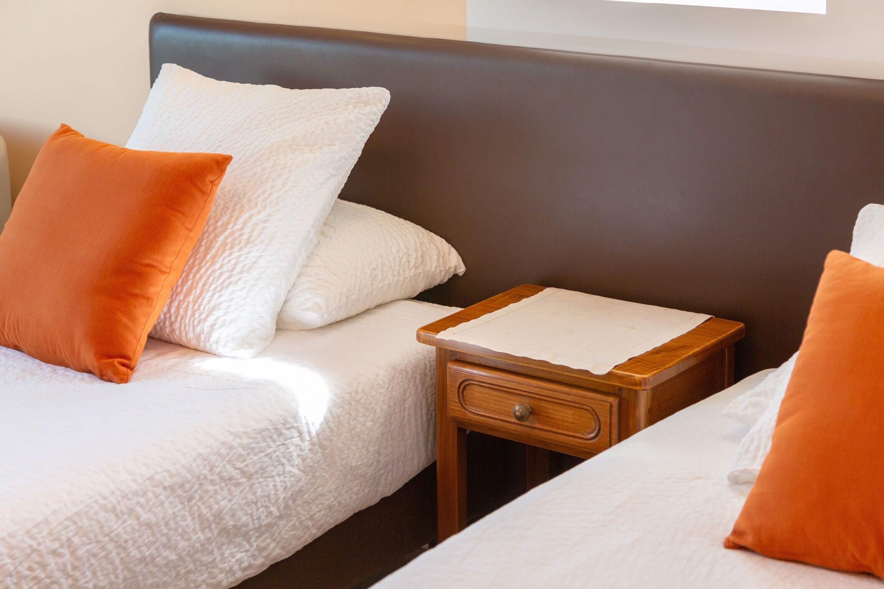 A hotel room with two beds, each with white bedding and orange pillows. There is a wooden nightstand with a drawer between the beds.