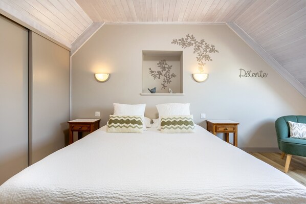 A bedroom with a large bed, two nightstands, a dresser, a chair, a wall-mounted shelf with decorative items, and a ceiling with wooden beams.