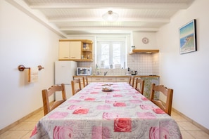 A dining area with a table covered in a floral tablecloth, wooden chairs, a kitchen in the background, and a window with a view.