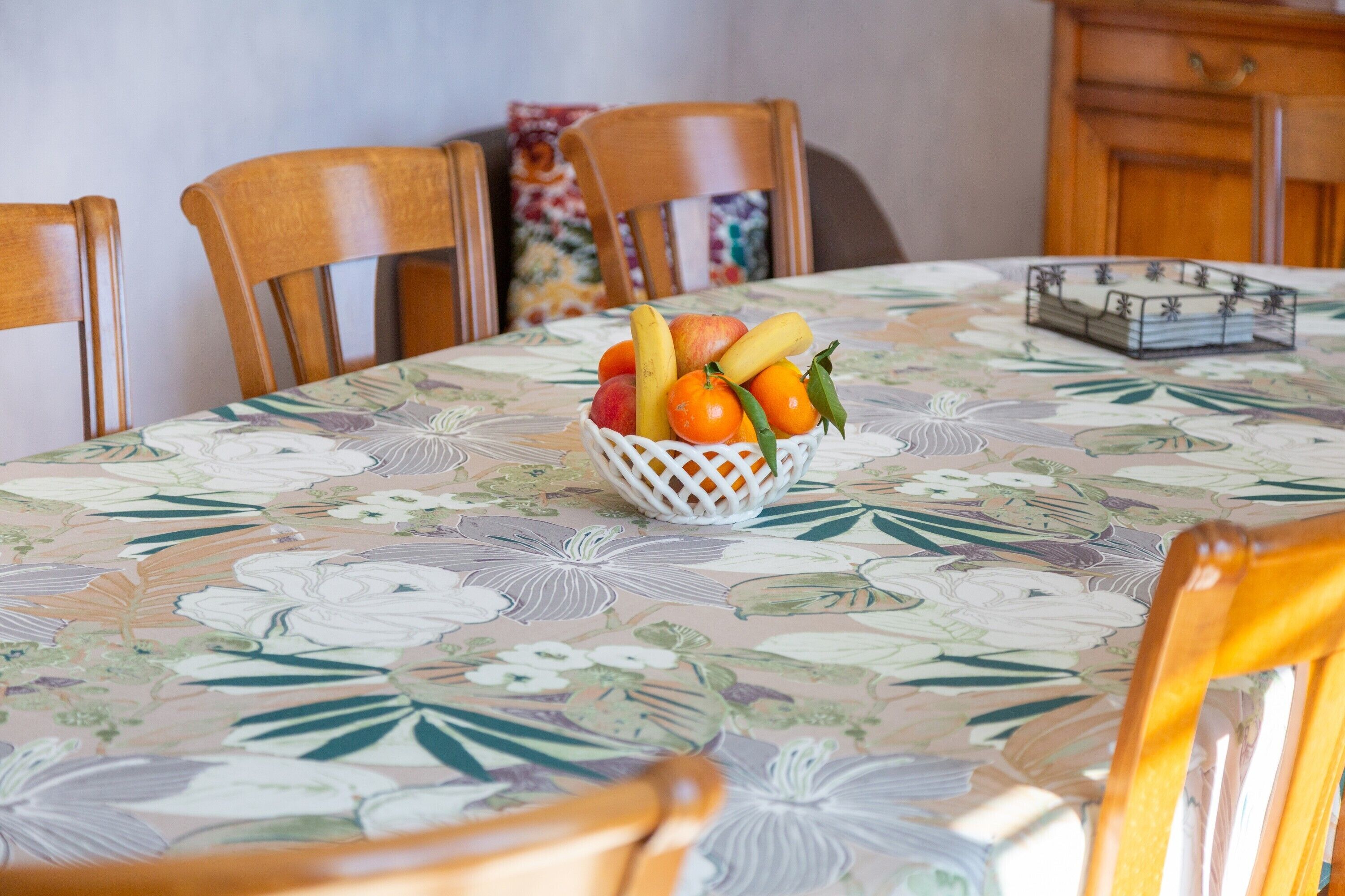 A dining table with a floral tablecloth, a fruit basket, and wooden chairs.