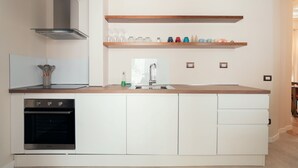 Une cuisine moderne avec des meubles blancs, un four en acier inoxydable, des étagères en bois au-dessus de l’évier et une fenêtre au-dessus de l’évier.