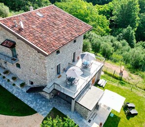 Un edificio de piedra con techo de tejas rojas, un patio cubierto con mobiliario exterior y un exuberante paisaje verde que lo rodea.
