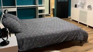 Une chambre à coucher comprenant un lit, une armoire, une table de chevet, un miroir et un petit ventilateur.