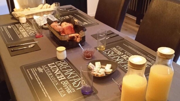 Une table de petit-déjeuner dressée avec un menu, du pain, des viennoiseries et deux bouteilles de jus d’orange.