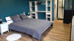 Une chambre à coucher comprenant un lit, une table de chevet blanche, un miroir, un tapis blanc, une armoire avec espace pour suspendre des vêtements et un espace douche.