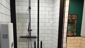 Un petit espace douche fermé, doté d’une porte en verre, de murs recouverts de carreaux blancs et de toilettes visibles à travers la porte.