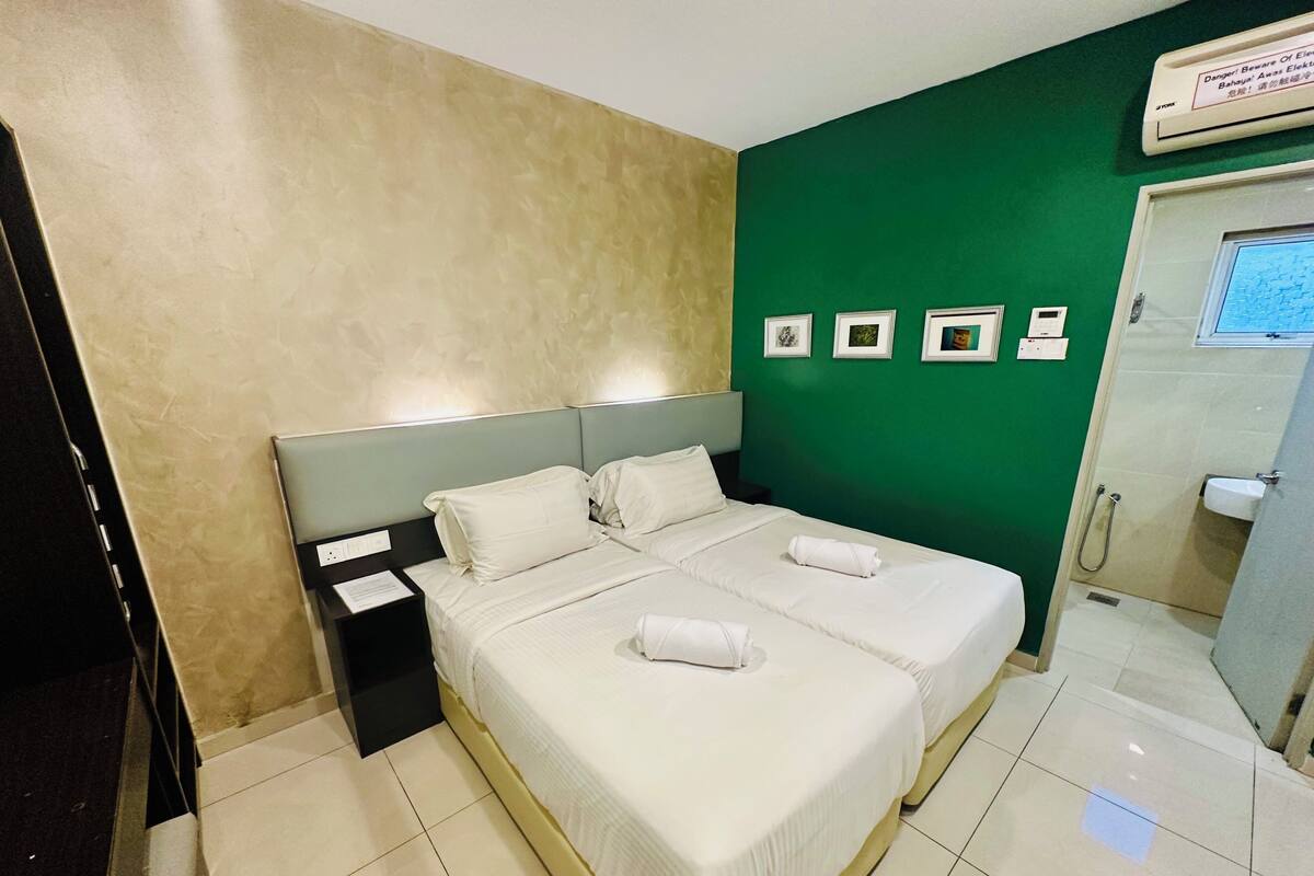 A hotel room with a double bed, a nightstand, a green accent wall, and a bathroom visible through an open door.