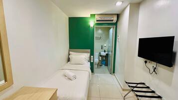 Quarto de hotel pequeno com cama, escrivaninha, televisão e banheiro visível através de uma porta aberta.