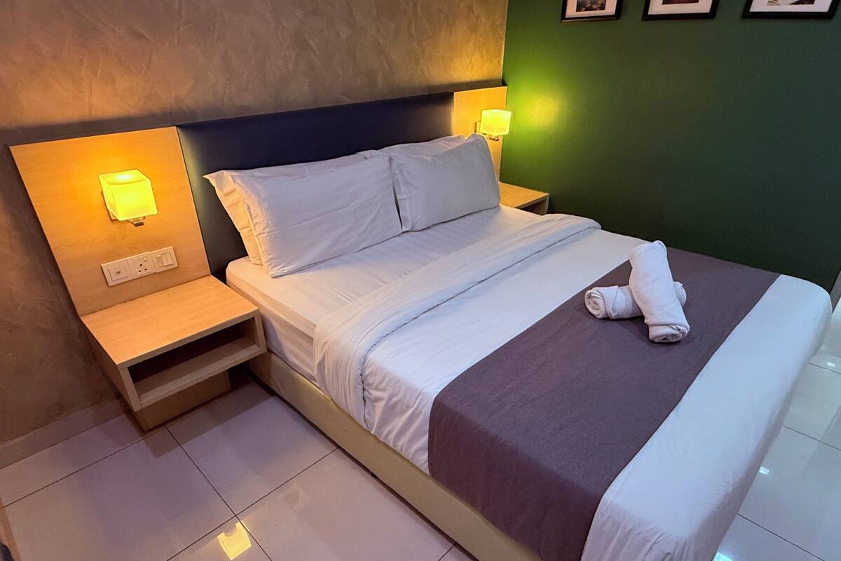 A neatly made bed with white linens and a folded towel on the side. There is a nightstand with a lamp and a power outlet. The room has a green wall and a textured ceiling.