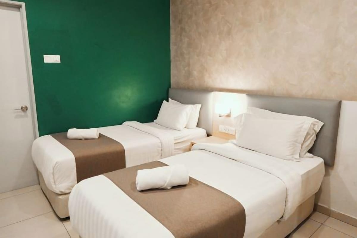 A hotel room with two beds, a green wall, and a nightstand with a lamp.