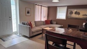 A living room with a sectional sofa, a dining table, and chairs. There is a window with curtains, a door, and a painting on the wall. - Elegant & Serene Urban Sanctuary (Halifax)