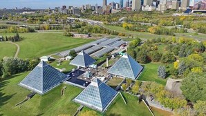 Een uitgestrekt zonnepark met veelhoekige zonnepanelen in een groen veld, omgeven door bomen en een stadsachtergrond.