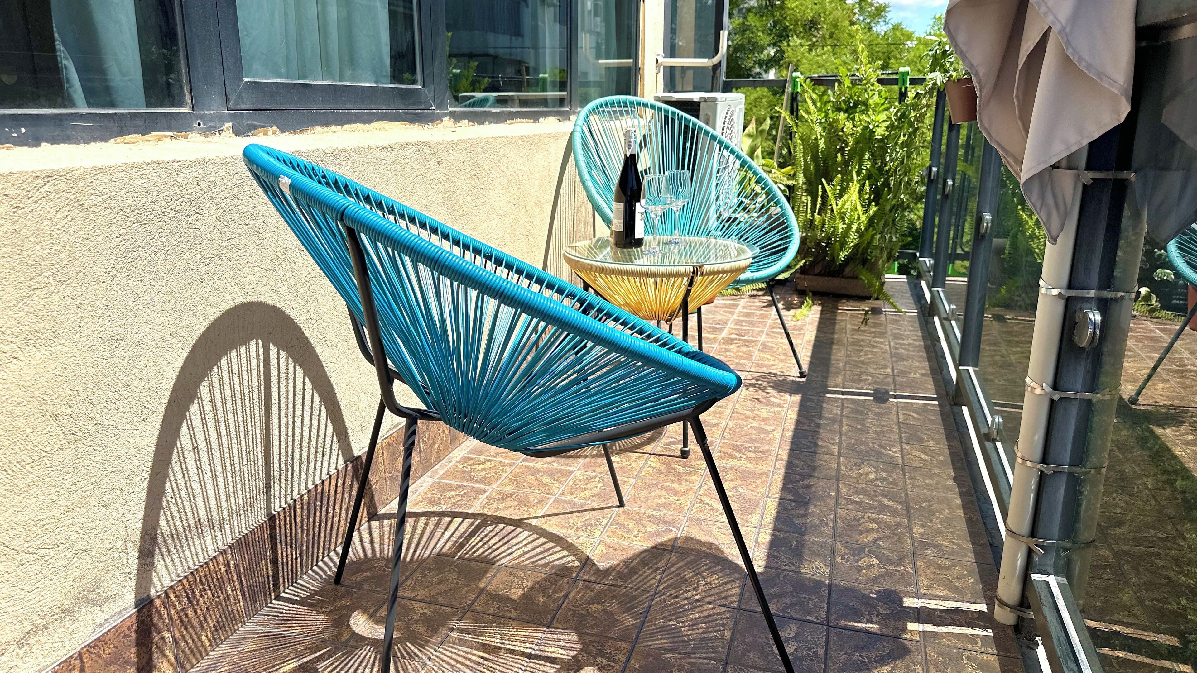 Two blue wicker chairs with a glass table, positioned on a tiled patio with a metal railing and greenery in the background.