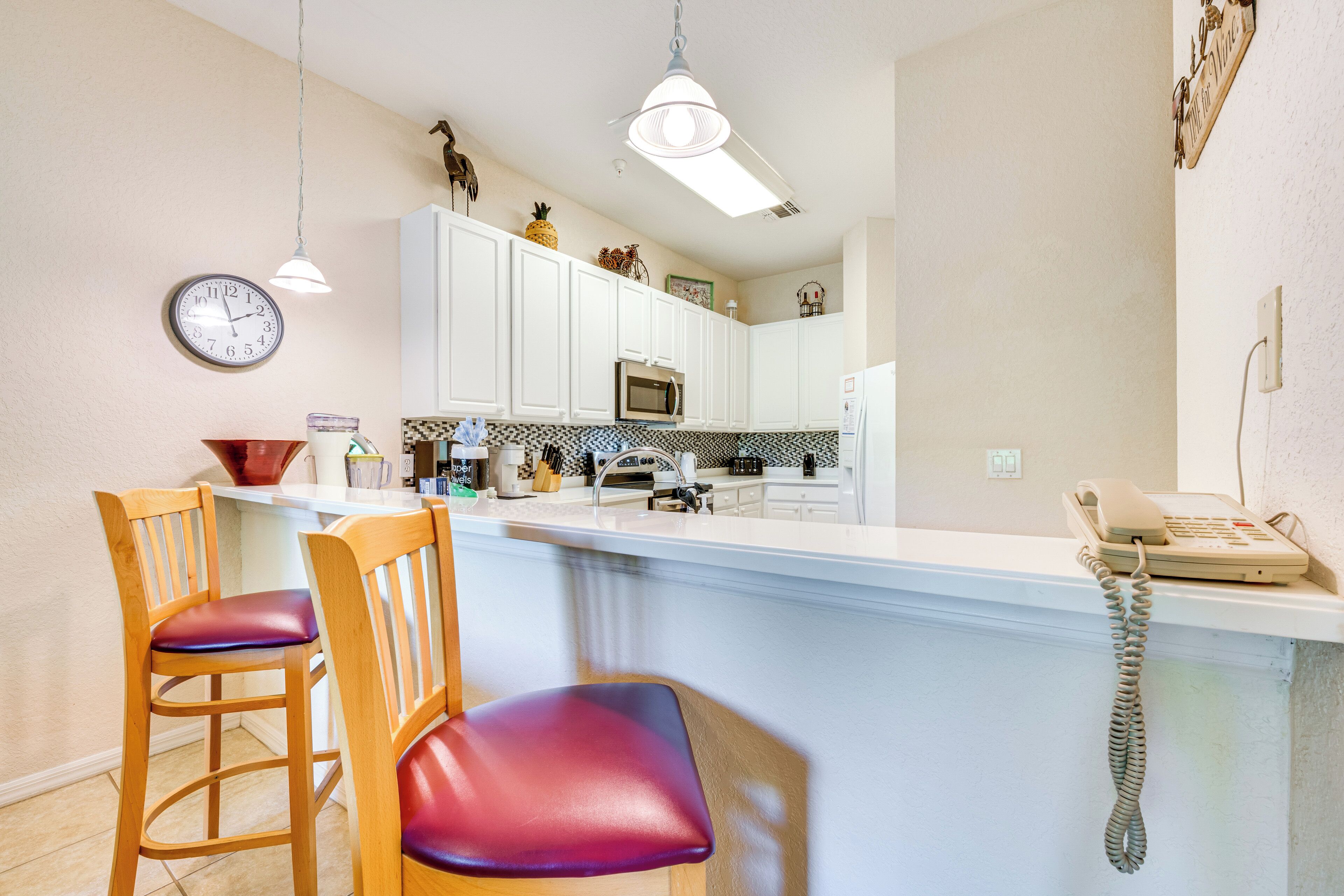 Een keuken met witte kasten, een magnetron, een koelkast en een telefoon op het aanrecht. Er staan twee houten stoelen met paarse kussens.