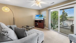 Een woonkamer met een plafondventilator, een flatscreen-tv, een bank, een bijzettafeltje en een balkon met uitzicht op de omgeving.
