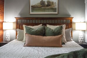 A neatly made bed with a quilted white cover, flanked by two bedside lamps with white bases and patterned lampshades, and a wooden headboard with a framed landscape painting above. - Hot Tub + Outdoor Kitchen: Stylish Home in Bryan (Bryan)