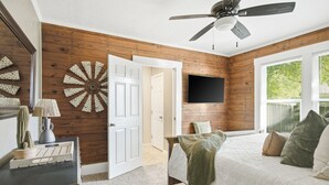 A bedroom with a wooden wall, a ceiling fan, a bed with pillows, a mounted TV, and a door leading to another room. - Hot Tub + Outdoor Kitchen: Stylish Home in Bryan (Bryan)