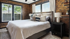 A bedroom with a large bed, wooden walls, and two windows with blinds. - Hot Tub + Outdoor Kitchen: Stylish Home in Bryan (Bryan)