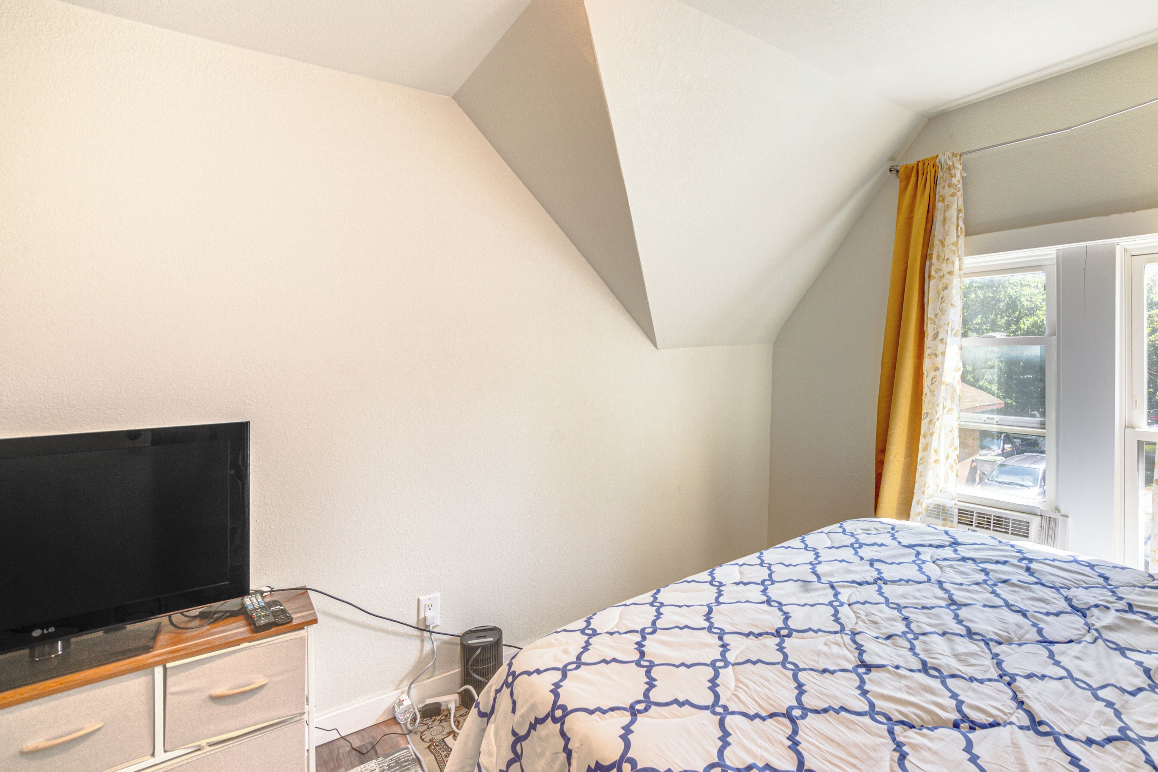 Ein Schlafzimmer mit Bett, Fernseher auf einem Holzschrank, einem Fenster mit Vorhängen und einer Heizung.
