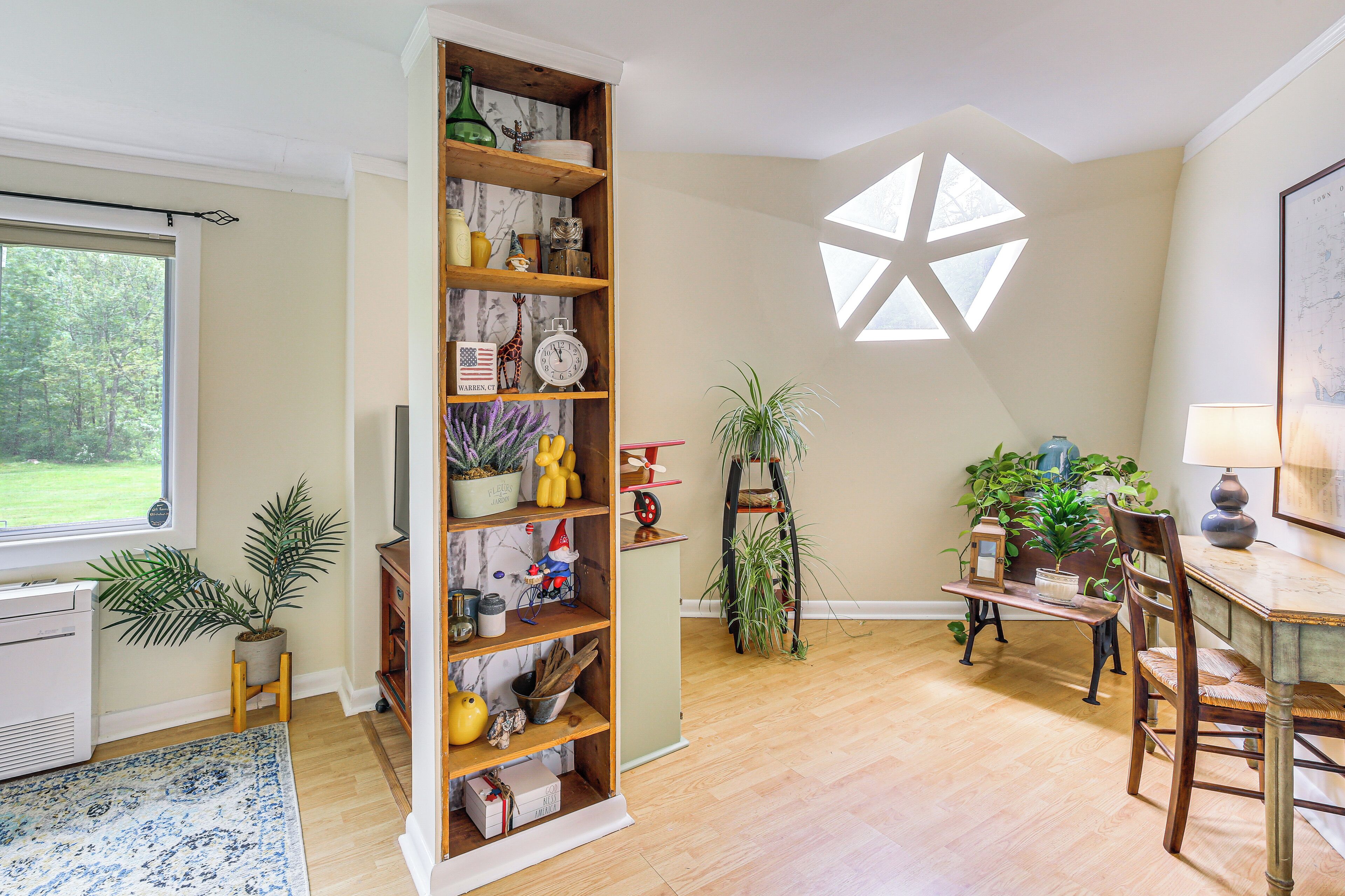 A room with a wooden bookshelf, a dining table with chairs, a lamp, and a large window with a view of trees.