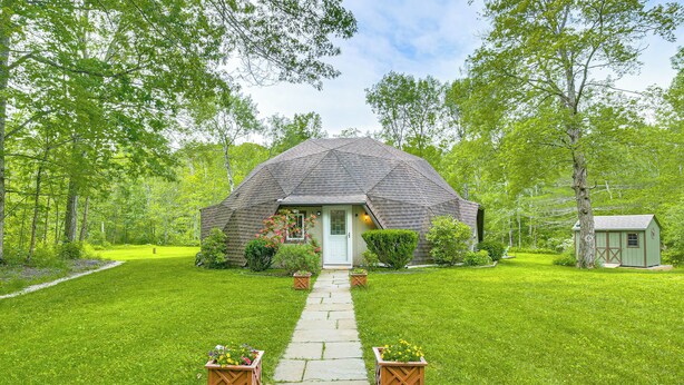 Une maison en forme de dôme géodésique, avec un chemin de pierre menant à l’entrée, entourée d’une végétation luxuriante et d’arbres.