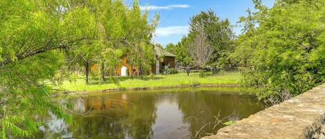 A rustic cabin with a pond in front, surrounded by lush greenery and a stone wall.