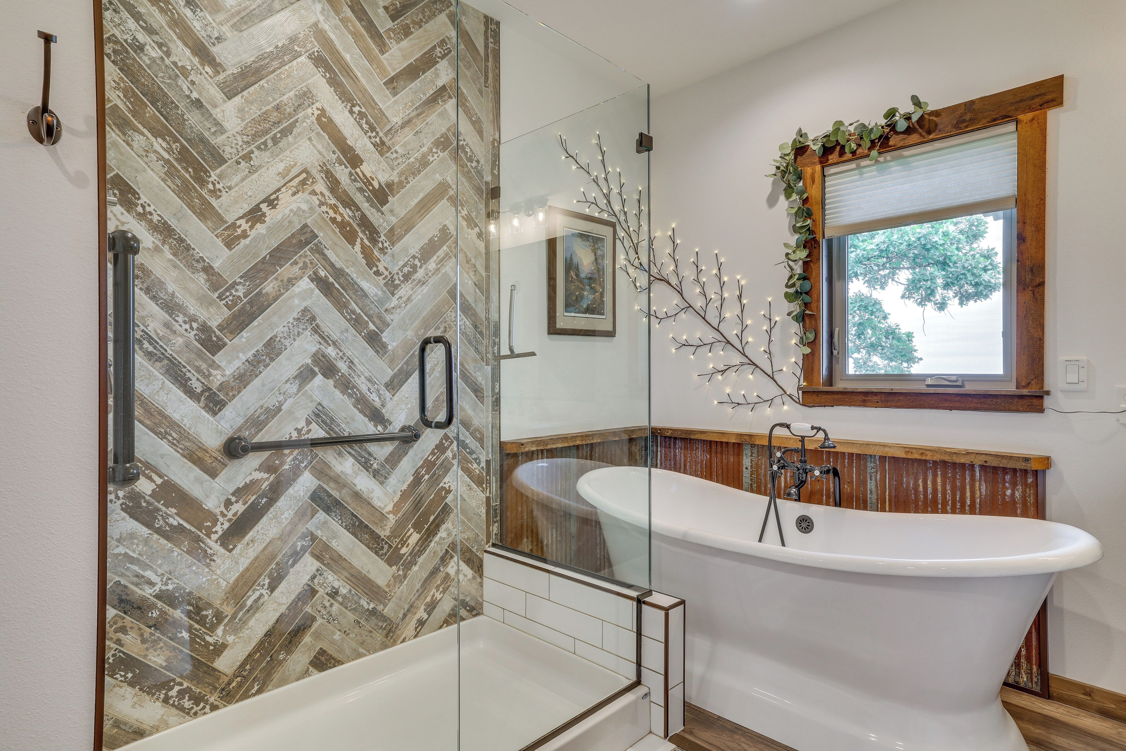 A bathroom with a herringbone-patterned wall, a freestanding bathtub, and a window with a wooden frame and greenery.