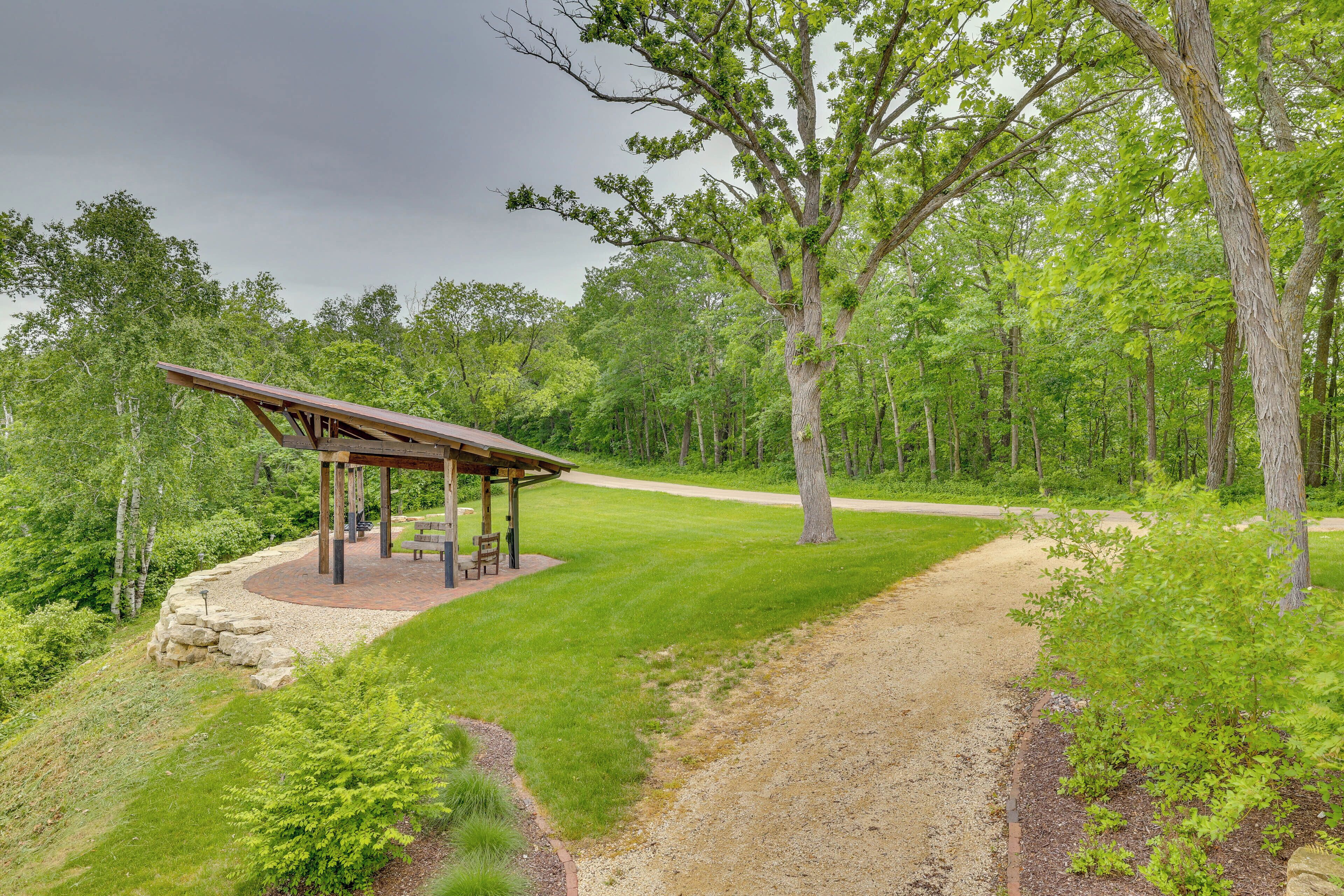 A covered pavilion with benches, surrounded by a gravel path and lush greenery.
