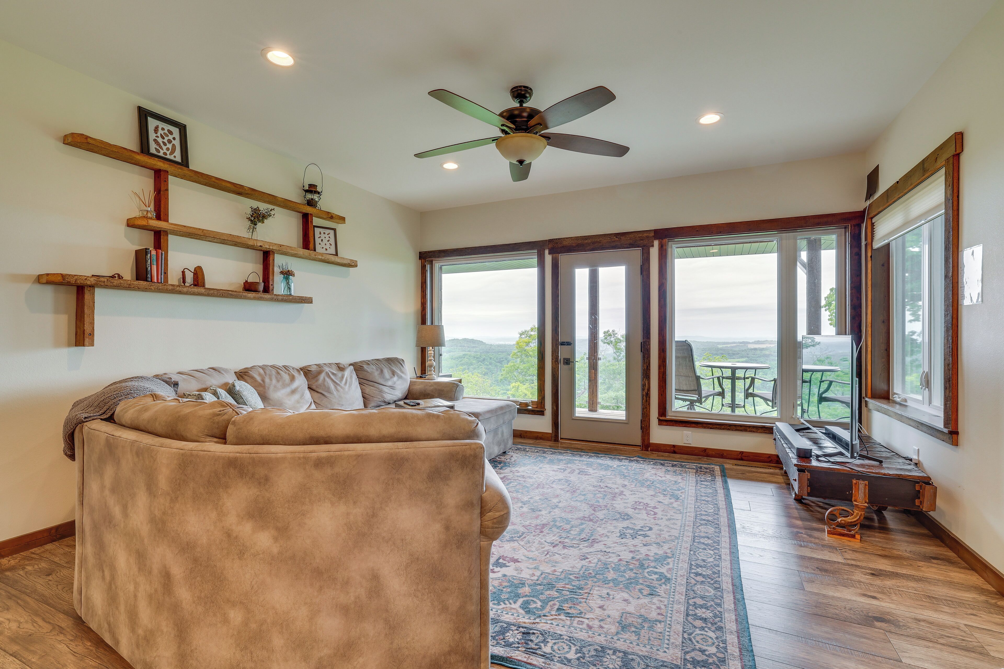 A spacious living room with a large beige sectional sofa, wooden shelves with decorative items, a ceiling fan, and a view through large sliding glass doors to an outdoor patio.