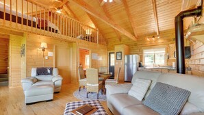 A spacious wooden cabin with a loft bedroom, a dining area, and a living room furnished with a sofa, armchair, and a checkered ottoman. - Bluff Views: 'bunker Hill' Cabin in Mindoro! (Mindoro)