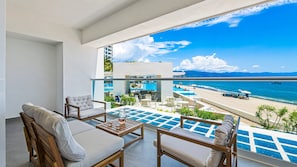 Un balcón con vista a una playa y zona de piscina, que incluye mobiliario exterior y una piscina con azulejos azules.