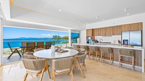 A modern kitchen with wooden cabinets and a large island, a dining area with wicker chairs, and a view of the ocean. - Beachfront, Private Pool, Theatre Room, Views! (Puerto Vallarta)