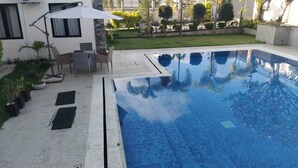 Una piscina con un borde de baldosas, una mesa y sillas, y una sombrilla blanca.