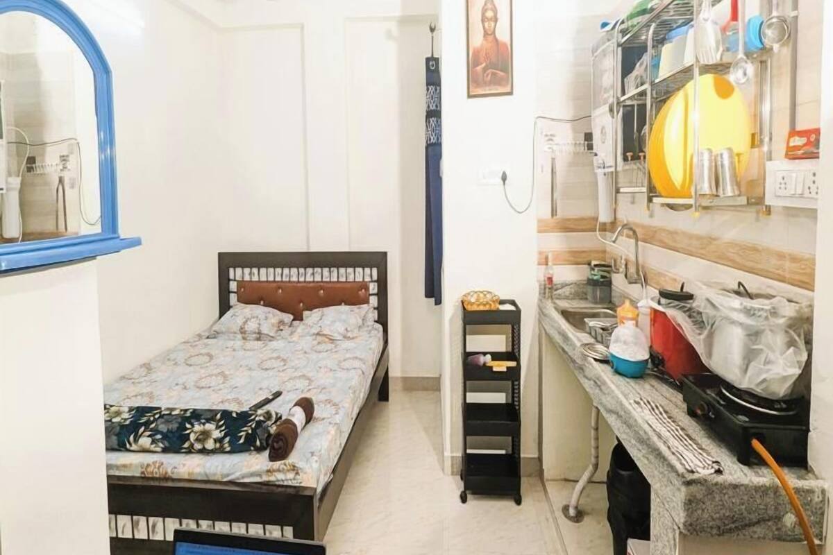 Une chambre à coucher de taille réduite, comprenant un lit simple, une petite table de chevet, un ventilateur de plafond et un coin cuisine avec un évier et divers ustensiles de cuisine.